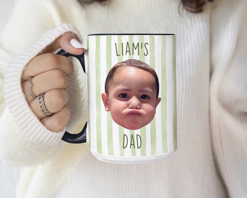 K&ouml;nnte beinhalten: Wei&szlig;er Keramikbecher mit schwarzem Henkel und Rand, mit einem gr&uuml;n-wei&szlig; gestreiften Muster. Der Becher hat ein Foto des Gesichts eines Kindes und den Text "LIAM'S DAD" in Gr&uuml;n.