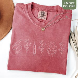 May include: A red t-shirt with a white design of a stethoscope, syringe, heart, and flowers. The shirt has the text "Comfort Colors" on the tag.