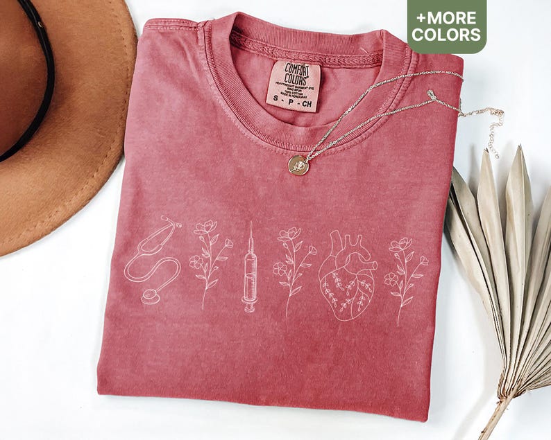 May include: A red t-shirt with a white design of a stethoscope, syringe, heart, and flowers. The shirt has the text "Comfort Colors" on the tag.