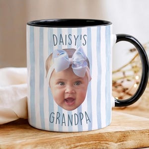 K&ouml;nnte beinhalten: Wei&szlig;er Keramikbecher mit schwarzem Innenraum und Griff. Der Becher zeigt das Gesicht eines Babys mit einer blauen Schleife und dem Text "DAISY'S GRANDPA" in Blau. Der Becher hat ein blau-wei&szlig; gestreiftes Muster.