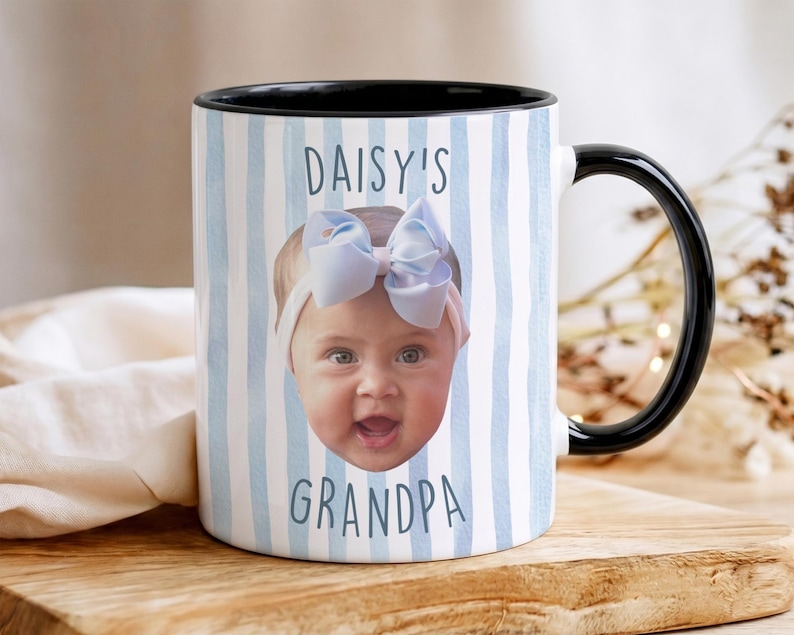 K&ouml;nnte beinhalten: Wei&szlig;er Keramikbecher mit schwarzem Innenraum und Griff. Der Becher zeigt das Gesicht eines Babys mit einer blauen Schleife und dem Text "DAISY'S GRANDPA" in Blau. Der Becher hat ein blau-wei&szlig; gestreiftes Muster.