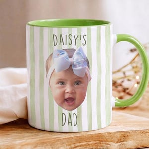 K&ouml;nnte beinhalten: Wei&szlig;er Keramikbecher mit gr&uuml;nem Henkel und Innenraum. Der Becher zeigt das Gesicht eines Babys mit einer blauen Schleife und dem Text "DAISY'S DAD" in Gr&uuml;n. Der Becher hat ein gr&uuml;n-wei&szlig;es Streifenmuster.