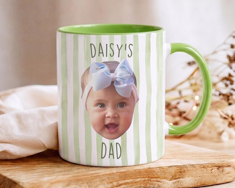 K&ouml;nnte beinhalten: Wei&szlig;er Keramikbecher mit gr&uuml;nem Henkel und Innenraum. Der Becher zeigt das Gesicht eines Babys mit einer blauen Schleife und dem Text "DAISY'S DAD" in Gr&uuml;n. Der Becher hat ein gr&uuml;n-wei&szlig;es Streifenmuster.