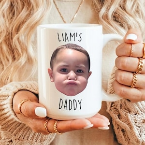 以下が含まれることがあります： 白いセラミックマグカップ。子供の顔と「LIAM'S DADDY」の文字が黒色でプリントされています。マグカップには大きなハンドルが付いています。マグカップを持っている人は金の指輪をしています。