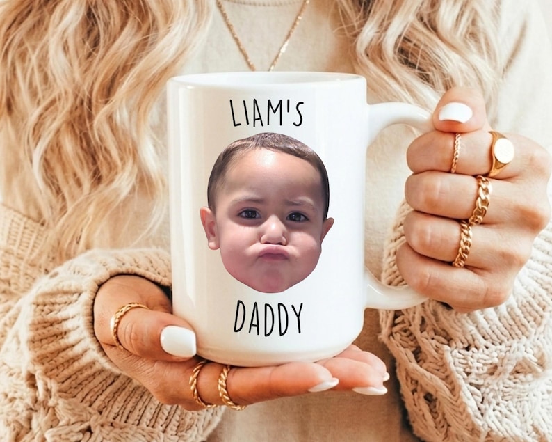 以下が含まれることがあります： 白いセラミックマグカップ。子供の顔と「LIAM'S DADDY」の文字が黒色でプリントされています。マグカップには大きなハンドルが付いています。マグカップを持っている人は金の指輪をしています。