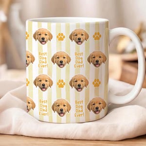 Może przedstawiać: Biały ceramiczny kubek z powtarzającym się wzorem pyszczk&oacute;w szczeniąt golden retriever, odcisk&oacute;w łap i napisem "Best Dog Dad Ever!" na ż&oacute;łto-białym tle w paski. Kubek ma biały uchwyt.