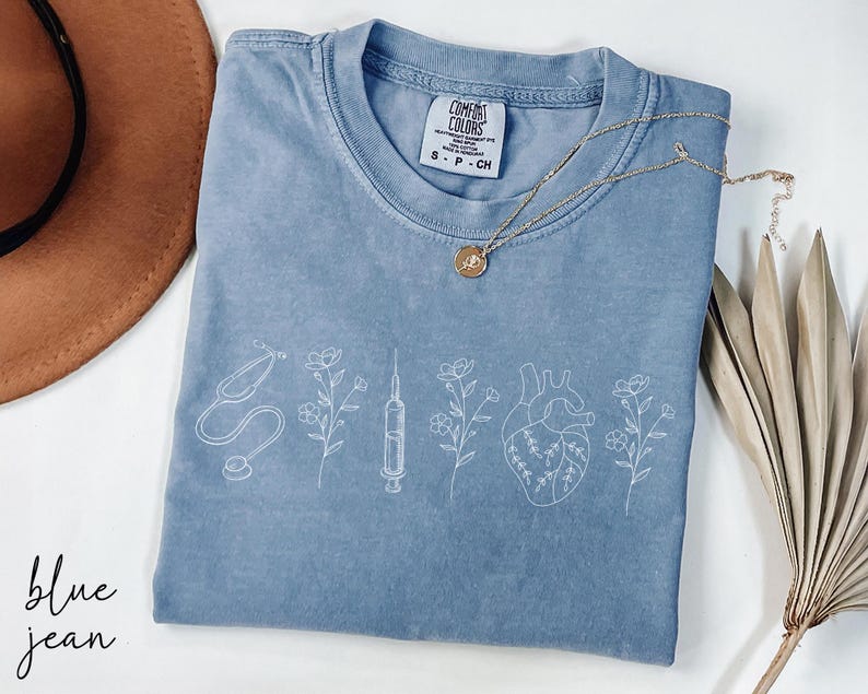 May include: A blue jean t-shirt with a white outline of a stethoscope, syringe, heart, and flowers. The shirt has the text "Comfort Colors" and "S - P - CH" printed on the front.