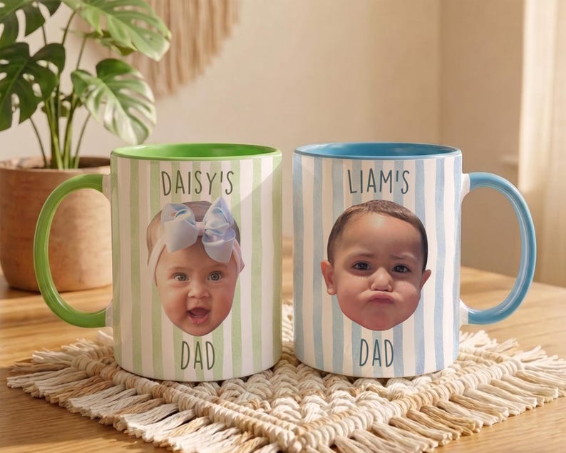 K&ouml;nnte beinhalten: Zwei Keramikbecher mit personalisierten Babyfotos. Ein Becher hat einen gr&uuml;nen Griff und Rand, vertikale gr&uuml;n-wei&szlig;e Streifen und den Text "DAISY'S DAD". Der andere Becher hat einen blauen Griff und Rand, vertikale blau-wei&szlig;e Streifen und den Text "LIAM'S DAD".