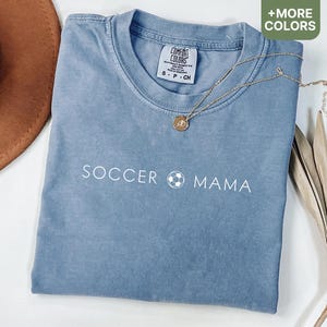 May include: A light blue t-shirt with the text "Soccer Mama" and a soccer ball graphic printed in white. The shirt is made of a soft, comfortable material.