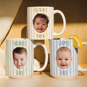 K&ouml;nnte beinhalten: Drei wei&szlig;e Keramikbecher mit individuellen Designs. Jeder Becher zeigt das Gesicht und den Namen eines Babys, mit den W&ouml;rtern "PAPA" und "DAD" darunter. Die Becher haben vertikale Streifenmuster in Gelb, Gr&uuml;n und Blau.