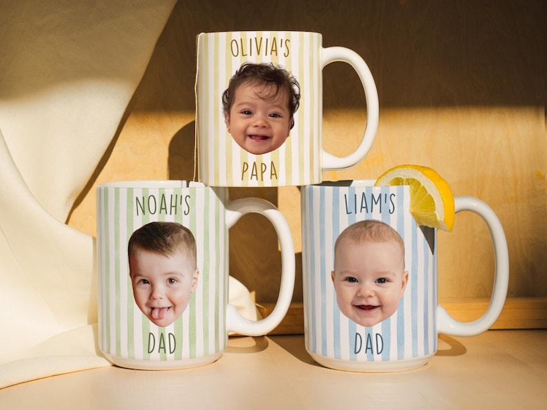 K&ouml;nnte beinhalten: Drei wei&szlig;e Keramikbecher mit individuellen Designs. Jeder Becher zeigt das Gesicht und den Namen eines Babys, mit den W&ouml;rtern "PAPA" und "DAD" darunter. Die Becher haben vertikale Streifenmuster in Gelb, Gr&uuml;n und Blau.