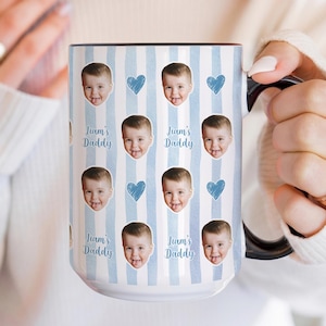 Może przedstawiać: Biały ceramiczny kubek z czarnym uchwytem i obramowaniem, z powtarzającym się wzorem twarzy dziecka, niebieskich serc i napisem "Liam's Daddy" na jasnoniebieskim tle w paski. Spersonalizowany prezent.