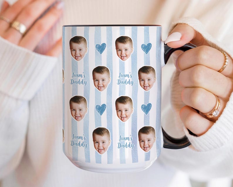 Może przedstawiać: Biały ceramiczny kubek z czarnym uchwytem i obramowaniem, z powtarzającym się wzorem twarzy dziecka, niebieskich serc i napisem "Liam's Daddy" na jasnoniebieskim tle w paski. Spersonalizowany prezent.