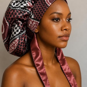 May include: A satin hair bonnet featuring a deep burgundy, black, and white geometric and circular pattern. The bonnet has a wide, smooth burgundy satin band and two long, matching burgundy satin ties that hang down from the front.