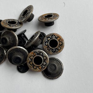 May include: A collection of circular metal rivets featuring a decorative floral-inspired embossed pattern on the top surface. The rivets have a weathered, dark silver-toned finish with visible oxidation and black stems extending from the back.