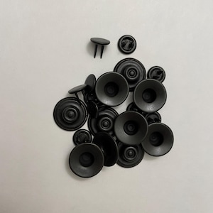 Op de afbeelding: Een verzameling zwarte metalen drukkers, inclusief zowel de mannelijke als de vrouwelijke componenten. De drukkers zijn in verschillende maten en vormen, met een matte afwerking. De afbeelding is van bovenaf genomen.