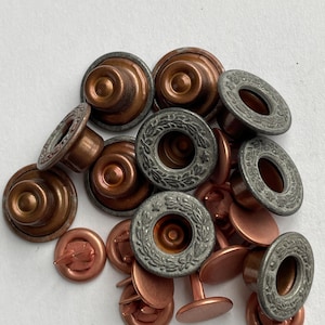 May include: A collection of metal buttons featuring a copper-toned base and a circular, antique silver-colored top rim decorated with an embossed laurel leaf pattern. These fasteners consist of two-part components with a central stud and a ring.