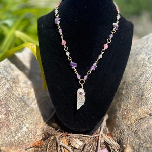 May include: A delicate necklace featuring a pendant made of a clear crystal wrapped in gold-colored wire. The necklace chain is adorned with small purple, pink, and clear gemstone beads. The necklace is displayed on a black mannequin.