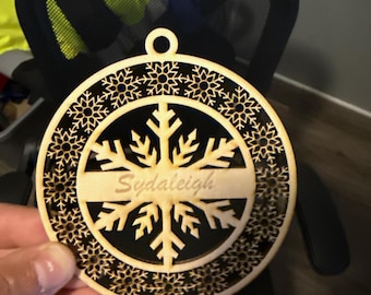 Op de afbeelding: Een lasergesneden houten kerstversiering met een sneeuwvlokontwerp. Het ornament heeft de naam "Sydaleigh" in een rechthoekige ruimte. De buitenrand is versierd met kleine sneeuwvlokpatronen. Er zit een gat aan de bovenkant om op te hangen.