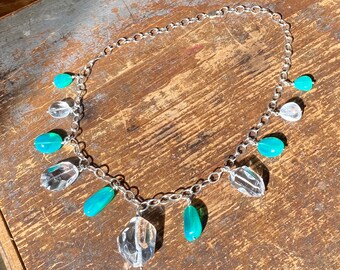 May include: A silver chain necklace with multiple teardrop-shaped clear and teal blue gemstone pendants.