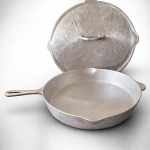 Puede incluir: Una sartén de hierro fundido de color plateado con tapa. La sartén tiene un mango largo y una superficie de cocción redonda. La tapa tiene un asa y diseños en forma de hojas. La batería de cocina es de metal.