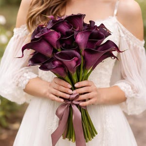 May include: A bouquet of deep purple calla lilies, tied with a mauve ribbon. The flowers have a velvety texture and are held against a white dress with off-the-shoulder sleeves. The stems are green and the overall arrangement is elegant.