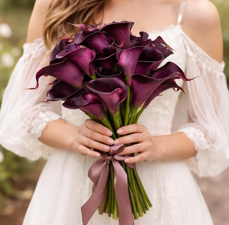 May include: A bouquet of deep purple calla lilies, tied with a mauve ribbon. The flowers have a velvety texture and are held against a white dress with off-the-shoulder sleeves. The stems are green and the overall arrangement is elegant.