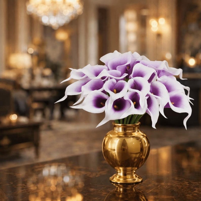 May include: A bouquet of purple and white calla lilies in a gold-colored vase. The flowers have a deep purple center and white petals. The vase is round with a pedestal base, and the background is blurred.