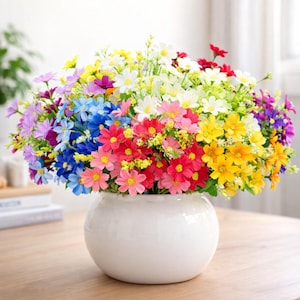 Op de afbeelding: Een levendig boeket van kleurrijke kunstbloemen in een witte keramische vaas. De bloemen zijn in tinten rood, roze, geel, blauw en paars. De vaas is rond en staat op een houten oppervlak.