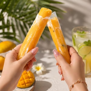 May include: Two orange mango-flavored ice pops in clear plastic sleeves held together. The frozen treats have a smooth, cylindrical shape with visible fruit pulp texture inside. The bright orange color contrasts with the transparent packaging.