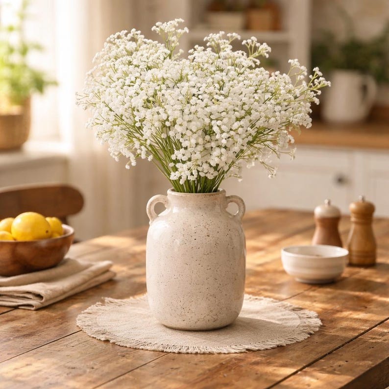 K&ouml;nnte beinhalten: Eine cremefarbene Vase mit zwei Henkeln enth&auml;lt einen Strau&szlig; wei&szlig;er Blumen. Die Vase steht auf einem runden, beigefarbenen Platzset auf einem Holztisch. Eine Holzschale mit Zitronen und Salz- und Pfefferstreuer stehen ebenfalls auf dem Tisch.