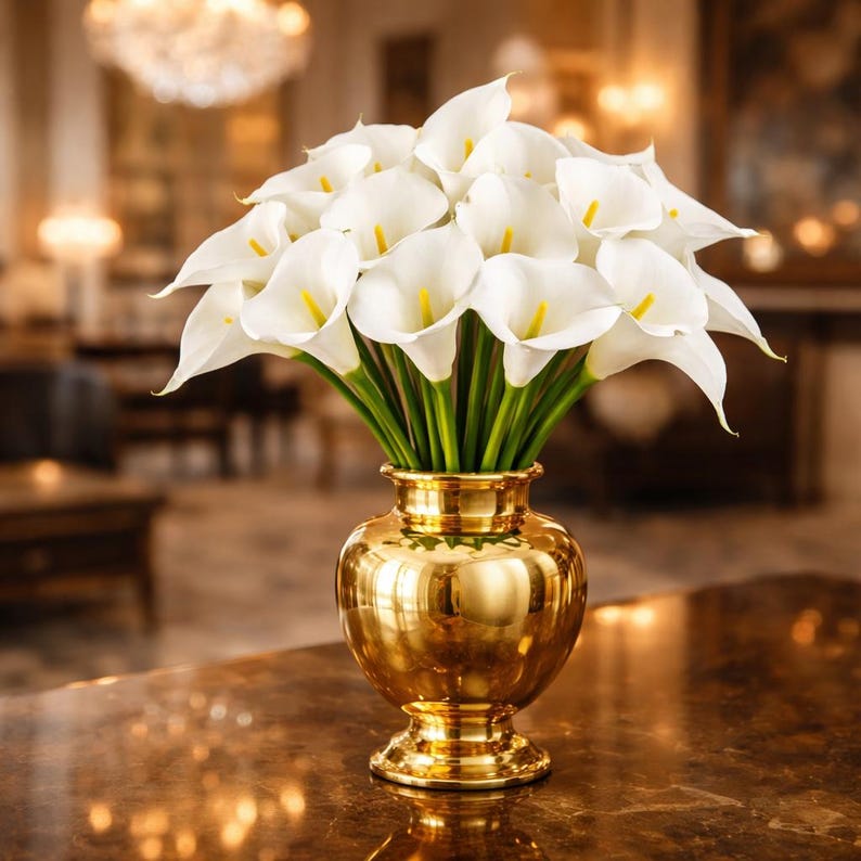May include: A bouquet of white calla lilies with yellow centers in a shiny gold vase. The vase has a bulbous body and a pedestal base. The flowers have green stems and are arranged to create a full, elegant display.
