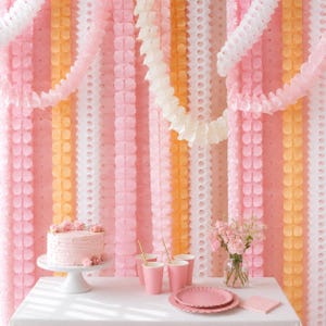 May include: A light pink frosted cake on a white pedestal stand, three pink paper cups, a stack of pink plates, and a glass vase with pink roses. The background features vertical streamers in shades of white, light pink, and peach.