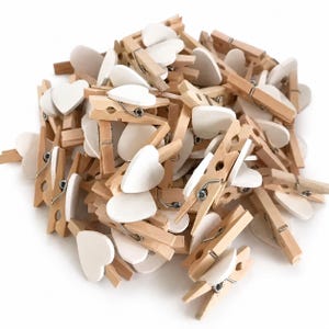 Könnte beinhalten: Ein großer Haufen kleiner Holzwäscheklammern, die jeweils mit einer flachen, weißen Herzform an der Oberseite versehen sind. Die Klammern bestehen aus hellem Naturholz und verfügen über einen silberfarbenen Metallfedermechanismus zum Öffnen.