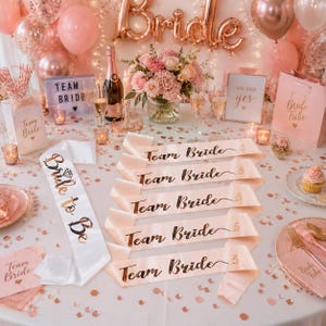 Op de afbeelding: Een set bruidsfeestaccessoires bestaande uit één witte satijnen sjerp met de tekst Bride to Be en vijf lichtroze satijnen sjerpen met de tekst Team Bride, uitgestald op een tafel met roségouden ballonnen, confetti en bloemendecoratie.