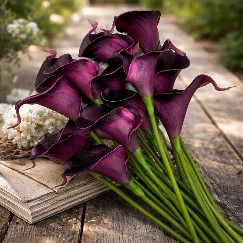 May include: A bouquet of deep purple calla lilies with long green stems. The flowers are arranged on a weathered wooden surface, with a few white flowers and a book visible in the background. The calla lilies have a trumpet-like shape.