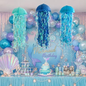 May include: A mermaid-themed birthday party display featuring three hanging jellyfish decorations in teal and blue, a tiered cake, and a backdrop with the text Mermaid Birthday, surrounded by blue, purple, and iridescent balloons and streamers.
