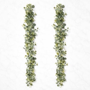 May include: Two vertical eucalyptus garlands with round, green leaves and small white flowers. The garlands are set against a white backdrop, offering a natural decorative touch. Suitable for home decor or event styling.