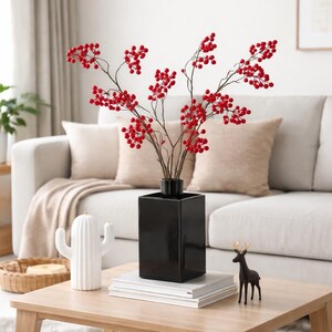 May include: A black, square vase displays branches laden with bright red berries. The vase is on a wooden coffee table, alongside a stack of books, a white cactus ornament, and a small black deer figurine. A beige sofa and cushions are in the background.