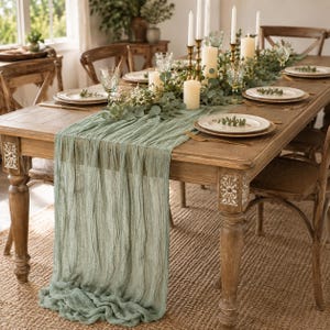 May include: A sage green cheesecloth table runner draped over a rustic wooden dining table. The table is set with white ceramic plates, gold-toned cutlery, clear glassware, and tall pillar candles nestled within a lush eucalyptus garland.