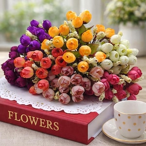 Op de afbeelding: Een kleurrijk boeket kunstbloemen in paars, oranje, rood, roze en wit. De bloemen liggen op een rood boek met gouden letters met de tekst "FLOWERS". Een witte kop met gouden stippen is ook zichtbaar.