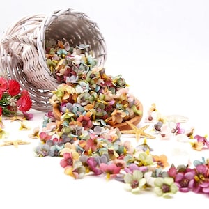 Könnte beinhalten: Ein weißer Weidenkorb, der überquillt mit einer Vielzahl kleiner, bunter Kunstblumen. Die Blumen sind in den Farben Rosa, Lila, Gelb und Grün. Der Korb ist umgekippt und die Blumen fallen auf eine weiße Oberfläche.