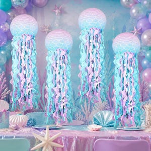 Puede incluir: Cuatro decoraciones colgantes de medusas con linternas de papel redondas con un patrón de escamas degradado en azul y morado, adornadas con largas serpentinas de papel onduladas en tonos azul claro, lavanda y morado que cuelgan hacia abajo.