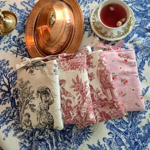 Könnte beinhalten: Vier kleine Stoffbeutel mit verschiedenen Mustern liegen auf einer blau-weiß geblümten Tischdecke. Die Beutel zeigen Toile-de-Jouy- und Blumenmuster in Rot, Rosa und Schwarz. Eine Kupferservierschale und eine Teetasse sind ebenfalls zu sehen.