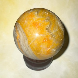 May include: A polished gemstone sphere with a warm color palette of yellow, orange, and gray. The orb is mounted on a dark brown wooden base. The sphere's surface has natural patterns and cracks.