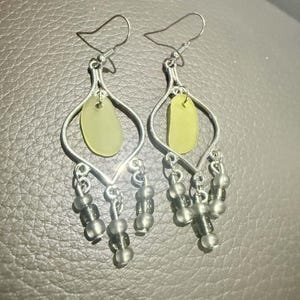 May include: A pair of silver-toned dangle earrings featuring a central pale yellow, smooth, rounded piece of sea glass suspended within a teardrop-shaped metal frame, accented with three small, translucent grey beads hanging from the base.