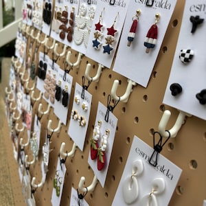 May include: A tan pegboard display featuring numerous pairs of earrings on white cards labeled esmeralda. The collection includes various shapes such as stars, popsicles, strawberries, flowers, and abstract white forms, all suspended by black hooks.