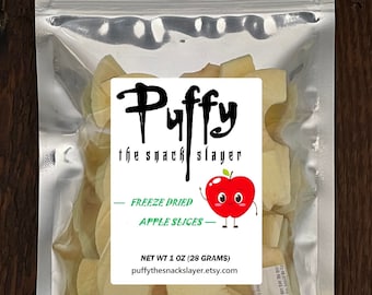 May include: A silver resealable bag filled with freeze-dried apple slices. The bag has a white label with the text "Puffy the snack slayer" and a cartoon apple with a smiling face. The label also says "Freeze Dried Apple Slices" and "Net Wt 1 oz (28 grams)". The website address "puffythesnackslayer.etsy.com" is printed on the label.