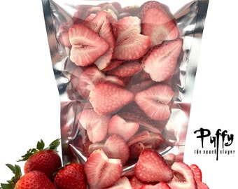 May include: A silver resealable bag filled with freeze-dried strawberry slices. Fresh strawberries are scattered on a white surface in front of the bag. The bag has a label that reads "Puffy the snack slayer".