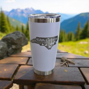 May include: A white stainless steel tumbler with a silver rim and lid. The tumbler features a gray outline of North Carolina with the text "North Carolina" and other graphics. The tumbler is sitting on a wooden table.